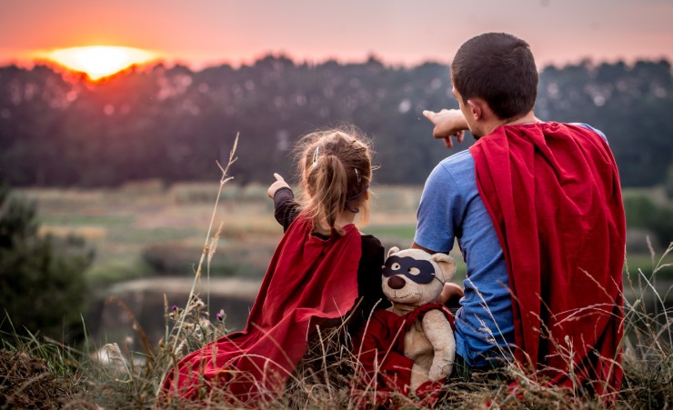 Father and daughter watching sun set in superman outfits.